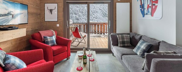 A cozy living room with a red armchair, a grey sofa, a wooden TV stand, and a balcony with a view of snow-covered trees.