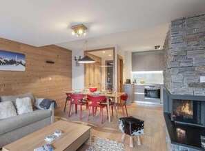 A modern living room with a fireplace, a dining area with red chairs, and a kitchen with stone accents.