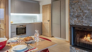 A modern kitchen with a stone fireplace, wooden cabinets, and a dining area with a table set for four. - 4-person flat - fireplace, close to the centre and slopes (Châtel)