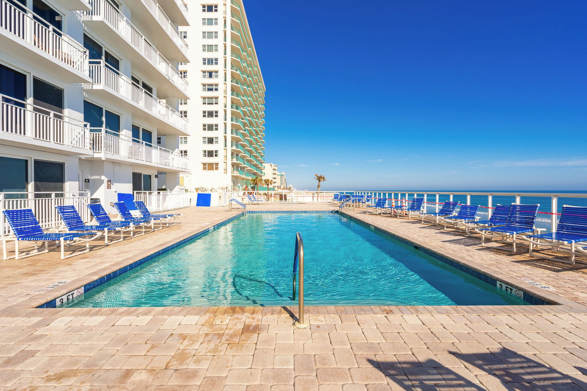 A swimming pool with blue lounge chairs on both sides, a clear blue sky, and a high-rise building in the background.