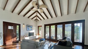 Ein geräumiges Wohnzimmer mit hoher Decke, einem großen Sofa, einem Fernseher und Blick ins Freie durch große Fenster.