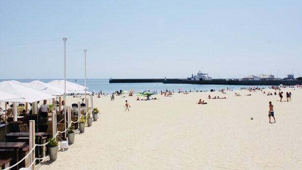 Ein Strand mit Sonnenschirmen und Tischen, ein Steg im Hintergrund und Personen im Sand.