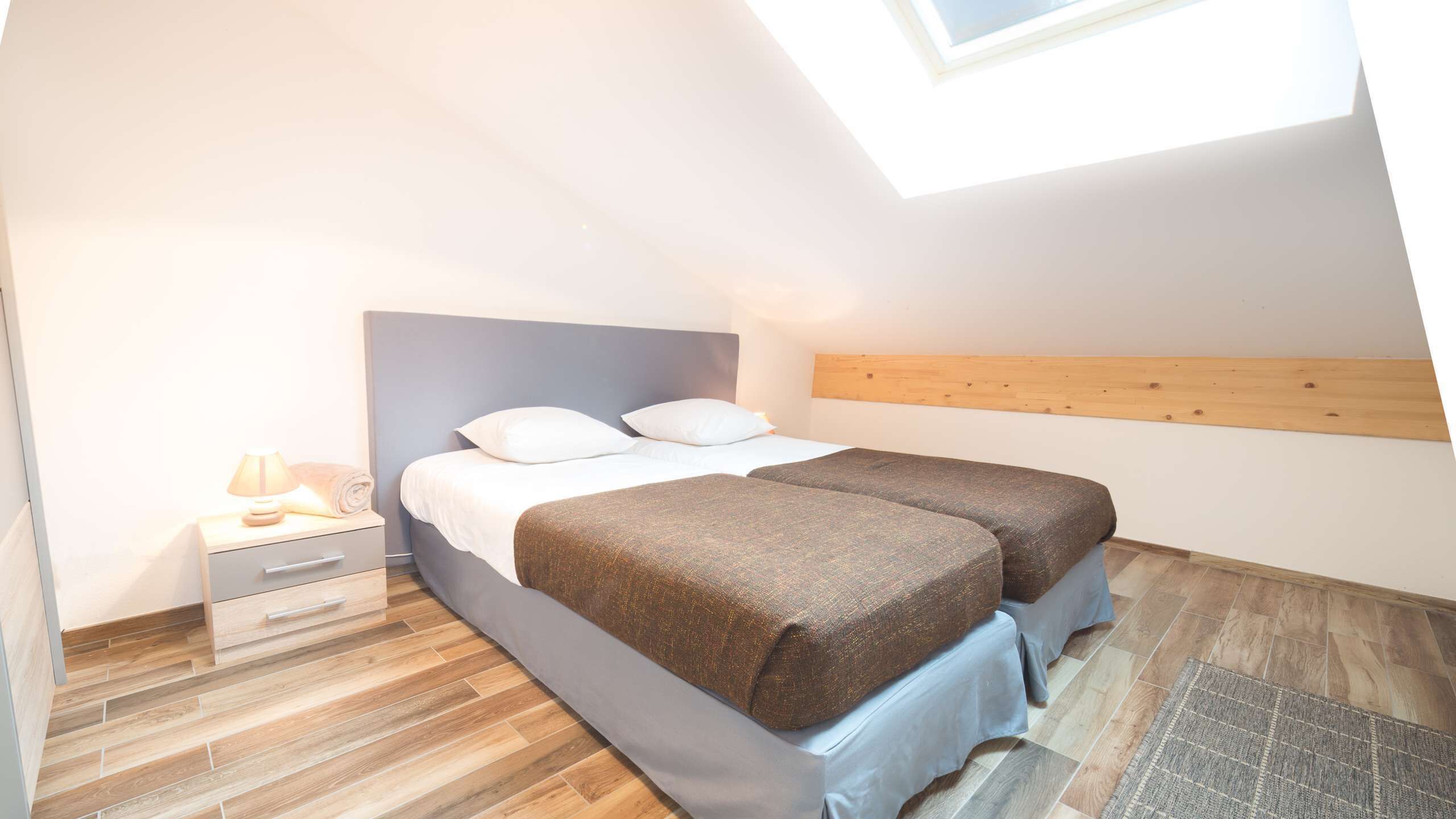 A small, modern bedroom with a skylight, a bed with two pillows, a bedside table with a lamp, and a wooden headboard.