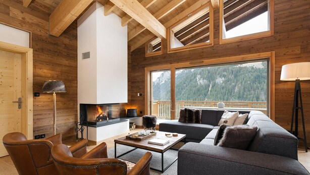 Ein gemütliches Wohnzimmer mit Kamin, Ledersesseln, einer Couch und einem großen Fenster mit Blick auf einen bewaldeten Berg.