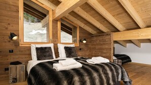 Ein Schlafzimmer mit Holzbalkendecke, einem Bett mit Felldecke, zwei Nachttischlampen und einem Fenster mit Blick auf eine verschneite Landschaft.