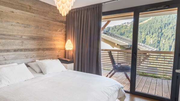 A bedroom with a wooden headboard, a large bed with white linens, a nightstand with a lamp, and a balcony with a view of a forested mountain.
