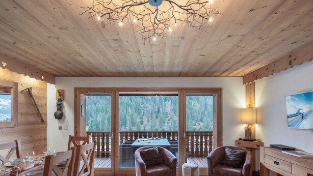 Interior acogedor de una cabaña con paredes de madera, una zona de comedor, dos sofás de cuero, una mesa de centro y un ventanal con vistas al bosque.