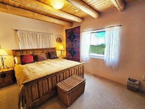 A bedroom with a wooden bed, a nightstand with a lamp, a wicker basket, a window with curtains, and a wall tapestry.