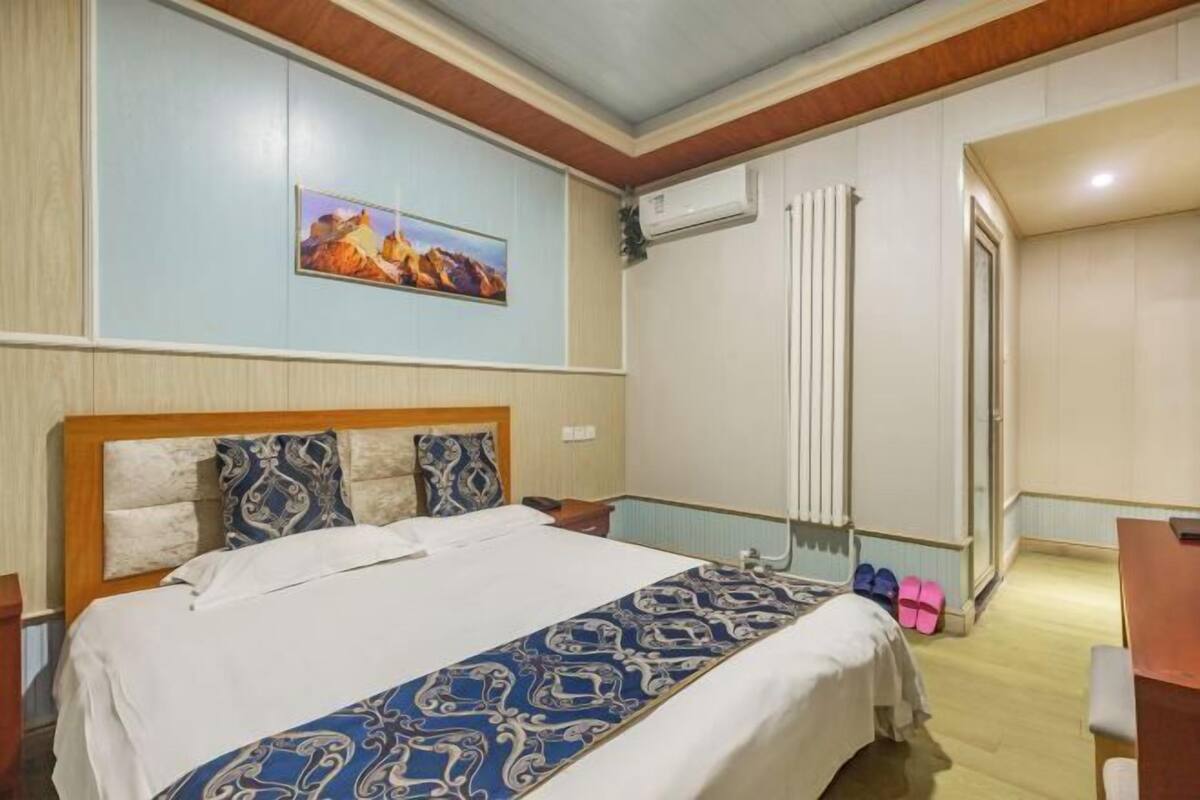 A hotel room with a bed, a painting on the wall, an air conditioning unit, and a door leading to another room.