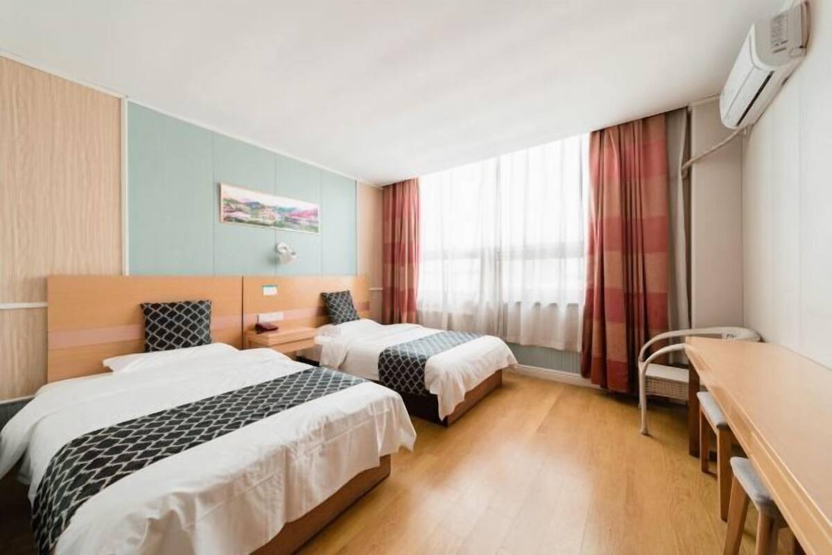 A hotel room with two beds, a desk, a chair, a window with curtains, and an air conditioning unit.
