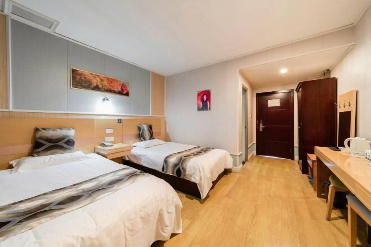 A hotel room with two beds, a desk, a chair, a painting on the wall, and a door leading to another room.