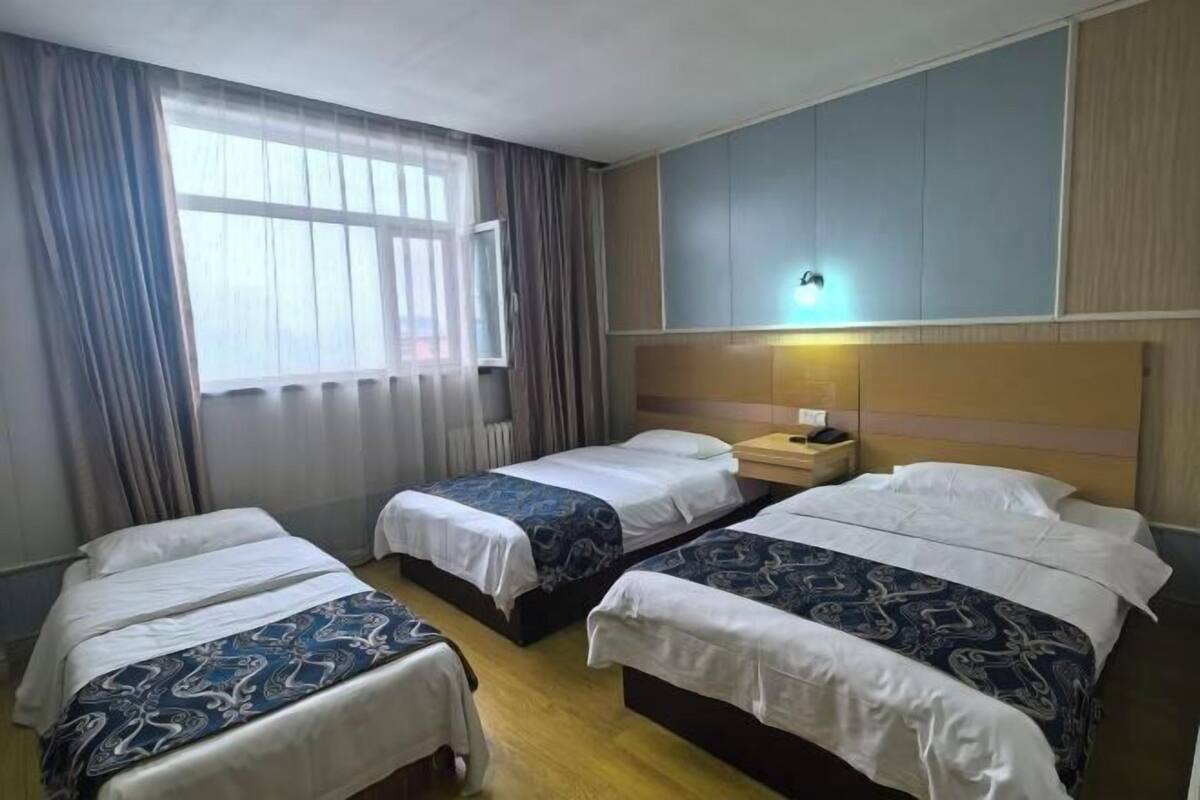 A hotel room with three beds, a window with curtains, a bedside table with a lamp, and a wall-mounted light fixture.