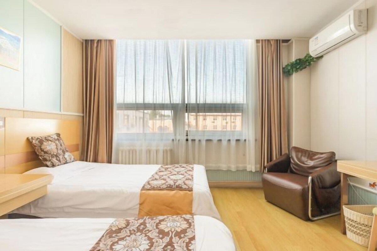 A hotel room with two beds, a desk, a leather chair, and a window with curtains.