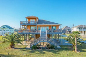 Una casa fronte spiaggia a più piani con un balcone, un pontile in legno e una scala che conduce all'ingresso.
