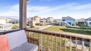 A residential area with houses, a grassy lawn, and a wooden deck with a chair. - Seaside MERRYment - 4 Bedroom + Bonus Room, 2 Bath, Sleeps 16. (Crystal Beach)