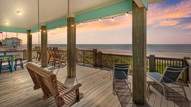 Une terrasse en bois avec des sièges d’extérieur, offrant une vue sur une plage au coucher du soleil.