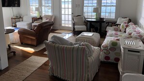 Ein geräumiges Wohnzimmer mit Holzboden, großen Fenstern mit Meerblick, einem braunen Ledersessel, einem gestreiften Sessel, einem weißen Sofa mit weißen Punkten und einem kleinen weißen Schrank.