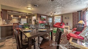 A spacious living area with a red sofa, dining table set with plates and glasses, and a kitchen with wooden cabinets and a ceiling fan. - License to Chill | Chill Mode Activated: Hot Tub, Pool Table & Cozy Fireplace (Ruidoso)