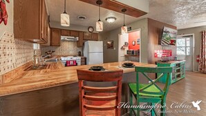 A kitchen with wooden cabinets, a central island, hanging pendant lights, and a view of a dining area with a green chair. - License to Chill | Chill Mode Activated: Hot Tub, Pool Table & Cozy Fireplace (Ruidoso)