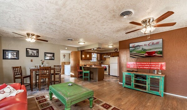 A spacious living room with a green coffee table, a red sofa, and a dining area with wooden chairs. There is a kitchen with wooden cabinets and a ceiling fan.