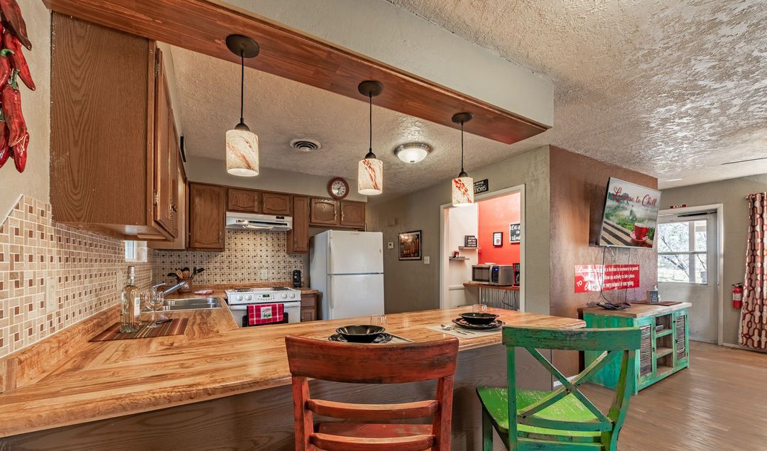 Una cucina con mobili in legno, un'isola centrale, lampade a sospensione e vista su una zona pranzo con una sedia verde.