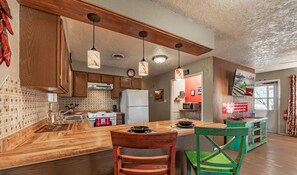 A kitchen with wooden cabinets, a central island, hanging pendant lights, and a view of a dining area with a green chair.