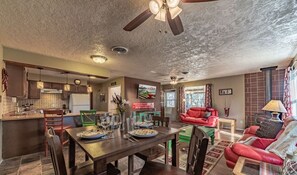 A spacious living area with a red sofa, dining table set with plates and glasses, and a kitchen with wooden cabinets and a ceiling fan.