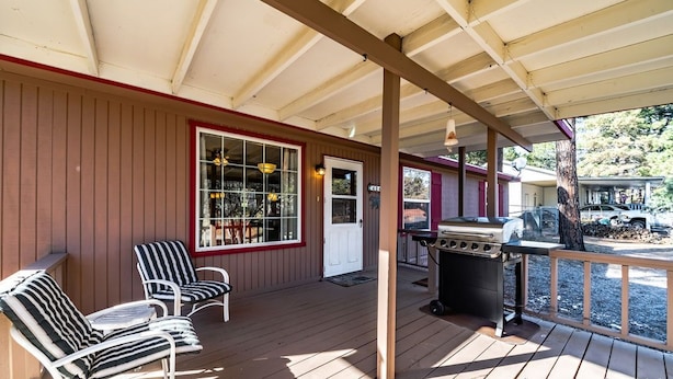 A covered porch with wooden flooring, two striped chairs, a grill, and a view of a vehicle in the background. - License to Chill | Chill Mode Activated: Hot Tub, Pool Table & Cozy Fireplace (Ruidoso)