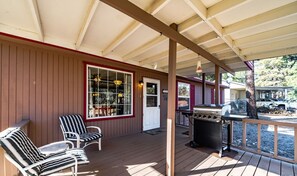 A covered porch with wooden flooring, two striped chairs, a grill, and a view of a vehicle in the background. - License to Chill | Chill Mode Activated: Hot Tub, Pool Table & Cozy Fireplace (Ruidoso)