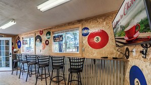 A bar area with pool tables and bar stools, a wooden wall with numbered targets, and a television screen. - License to Chill | Chill Mode Activated: Hot Tub, Pool Table & Cozy Fireplace (Ruidoso)