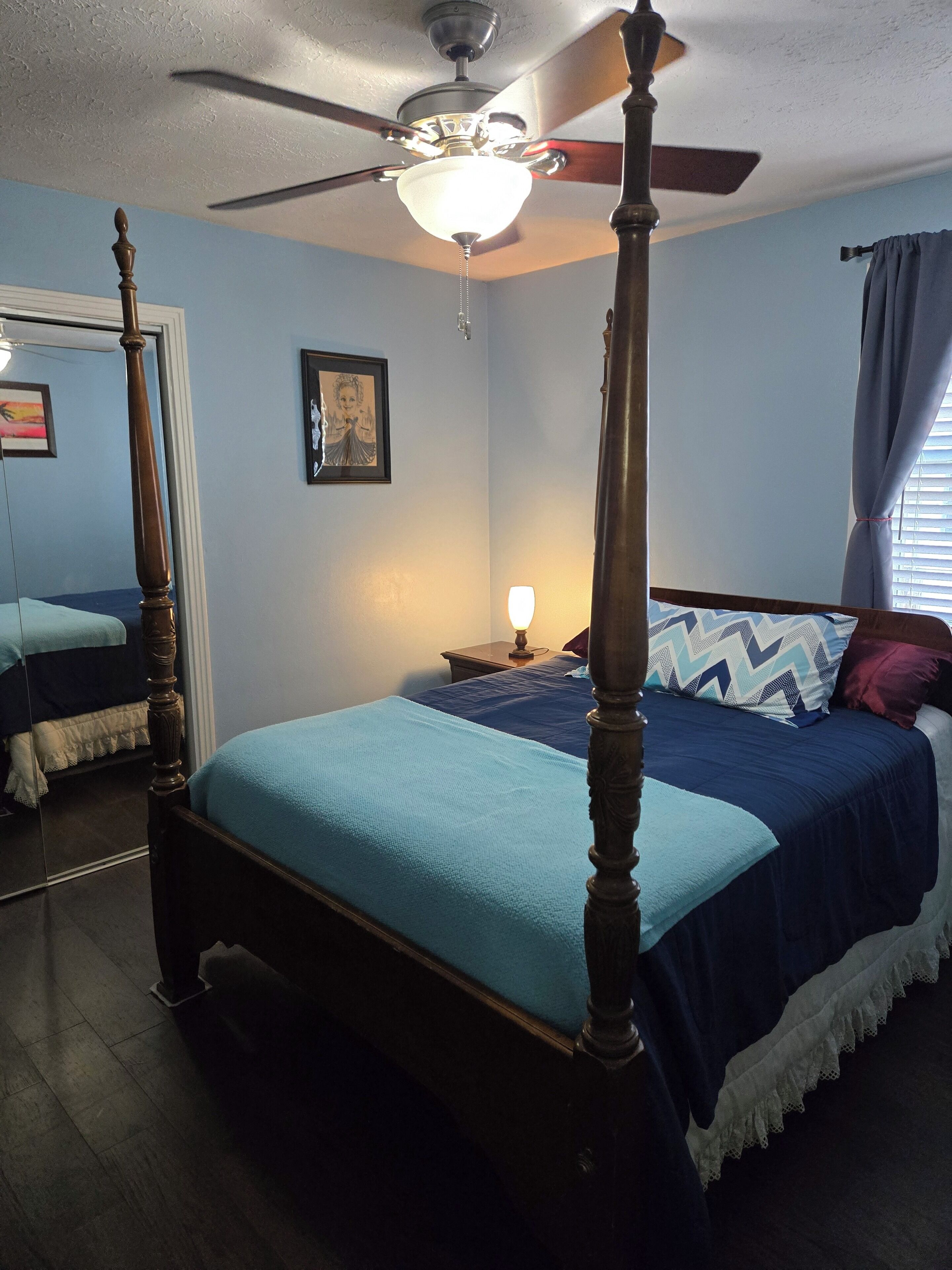 A four-poster bed with a blue bedspread, a nightstand with a lamp, a ceiling fan, and a framed picture on the wall.