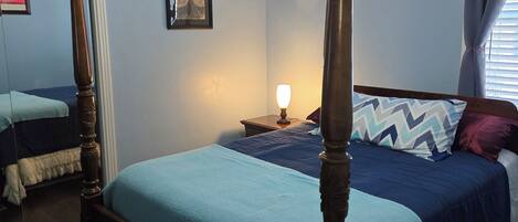 A four-poster bed with a blue bedspread, a nightstand with a lamp, a ceiling fan, and a framed picture on the wall.
