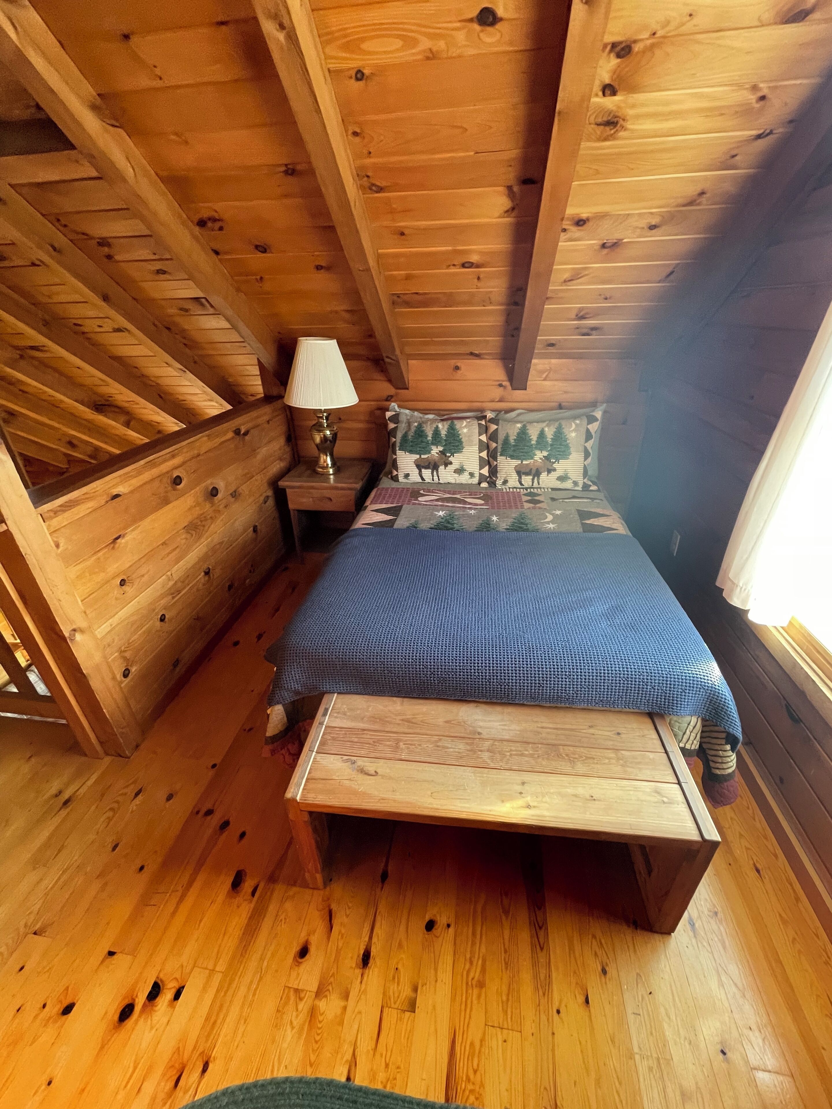 A wooden cabin bedroom with a bed, nightstand, lamp, and a window.