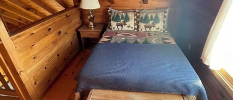 A wooden cabin bedroom with a bed, nightstand, lamp, and a window.