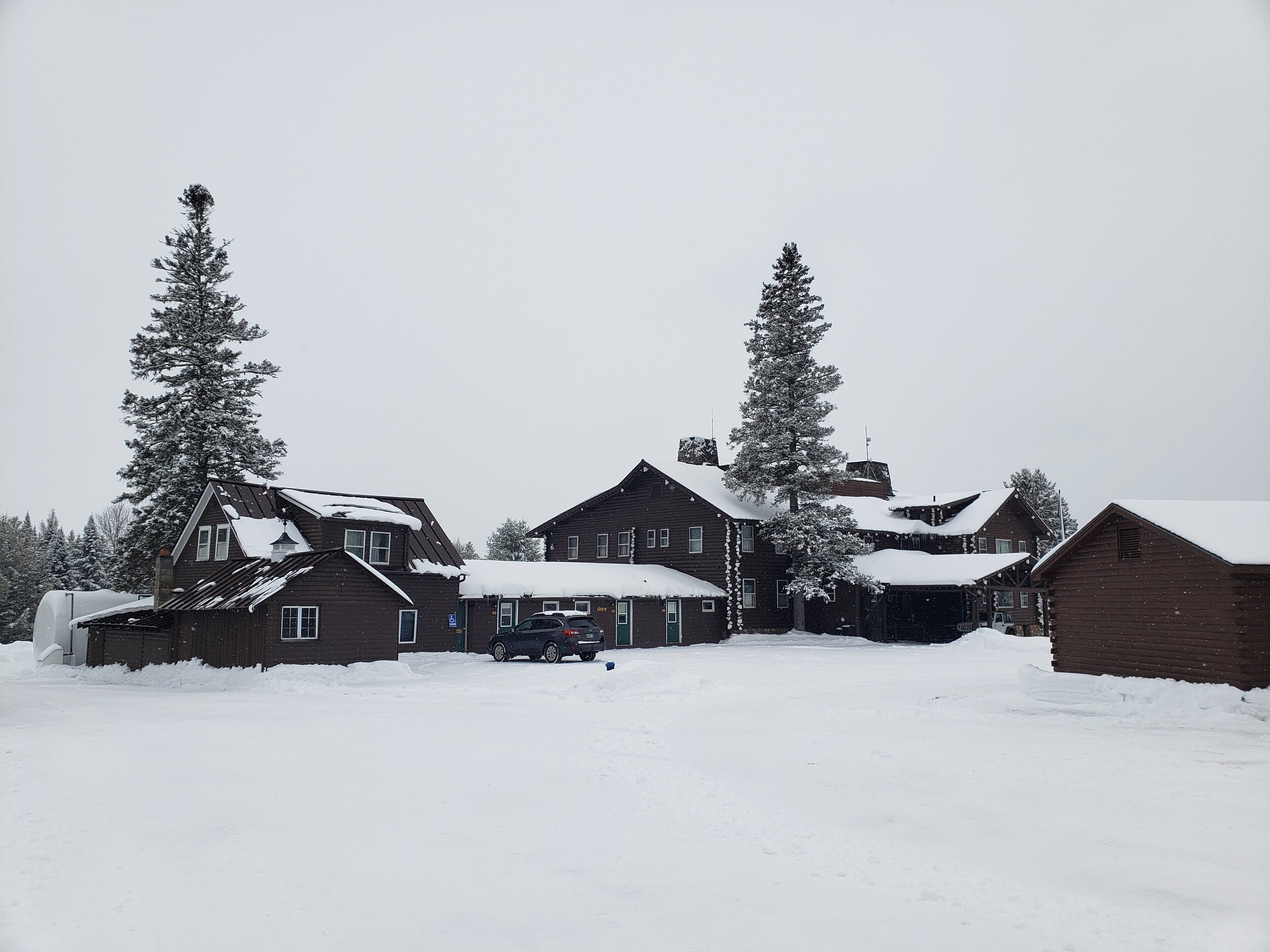 A snow-covered wooden cabin complex with multiple buildings, surrounded by tall evergreen trees.