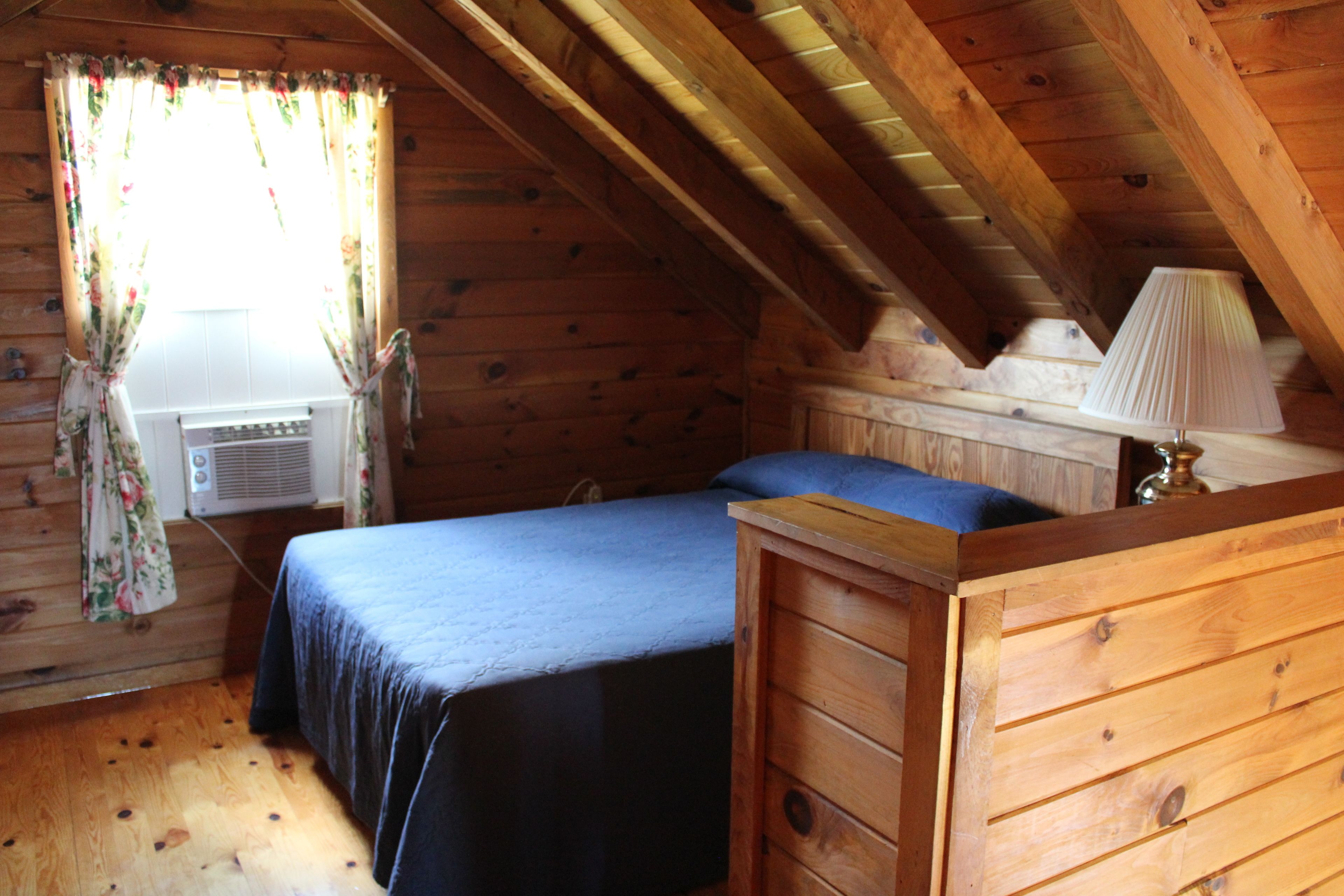 A wooden cabin bedroom with a bed, a dresser, a lamp, and an air conditioning unit.