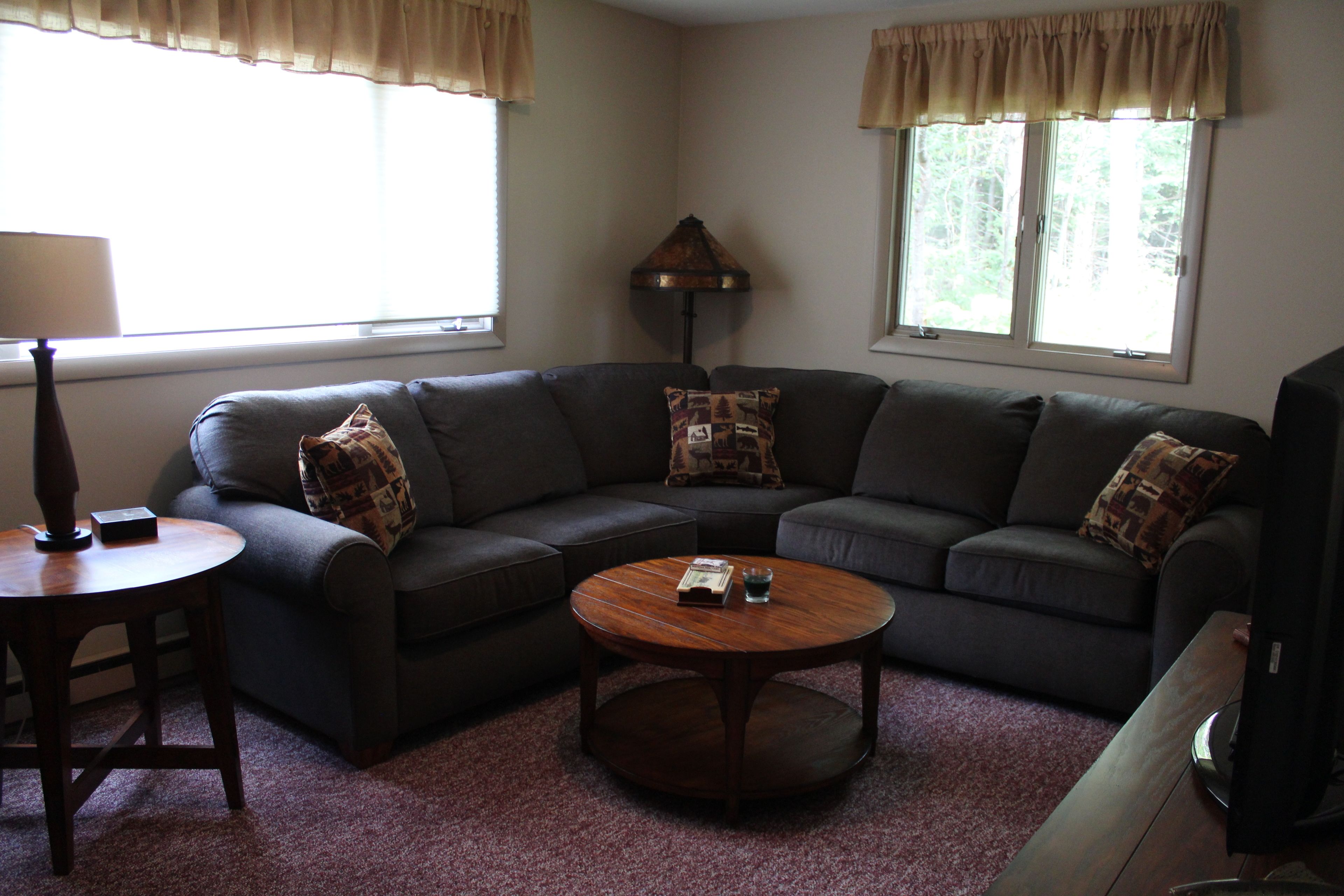 A living room with a dark-colored sofa, a round coffee table, a lamp, and two windows with curtains.