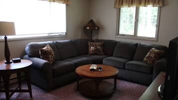 A living room with a dark-colored sofa, a round coffee table, a lamp, and two windows with curtains.