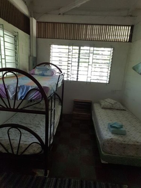 Una habitación pequeña y poco iluminada con una estructura de cama metálica, un colchón individual y una ventana con barrotes.