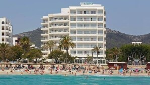 Ένα πολυώροφο ξενοδοχείο με μπαλκόνια, μια παραλία στο πρώτο πλάνο και βουνά στο βάθος.