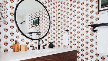 Une salle de bain avec un lavabo blanc, un meuble-lavabo en bois et un miroir. Le mur est décoré d’un motif répétitif représentant des visages de singes.