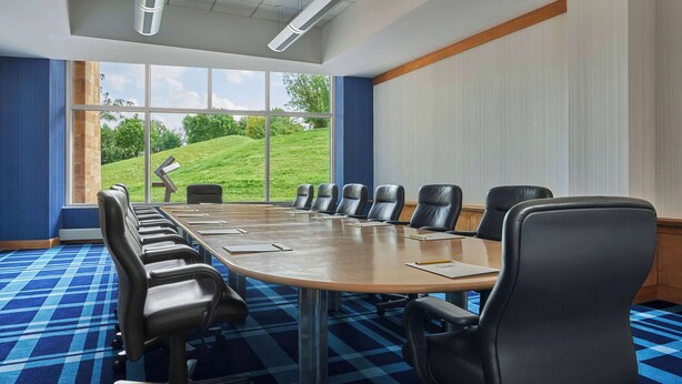 Un salón de conferencias con una larga mesa, múltiples sillas y ventanas amplias que dan a un paisaje cubierto de césped.