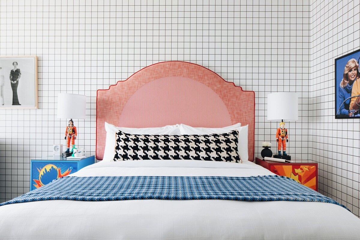 Una cama con colcha a cuadros, cabecera rosa y dos mesitas de noche con lámparas y figuritas.