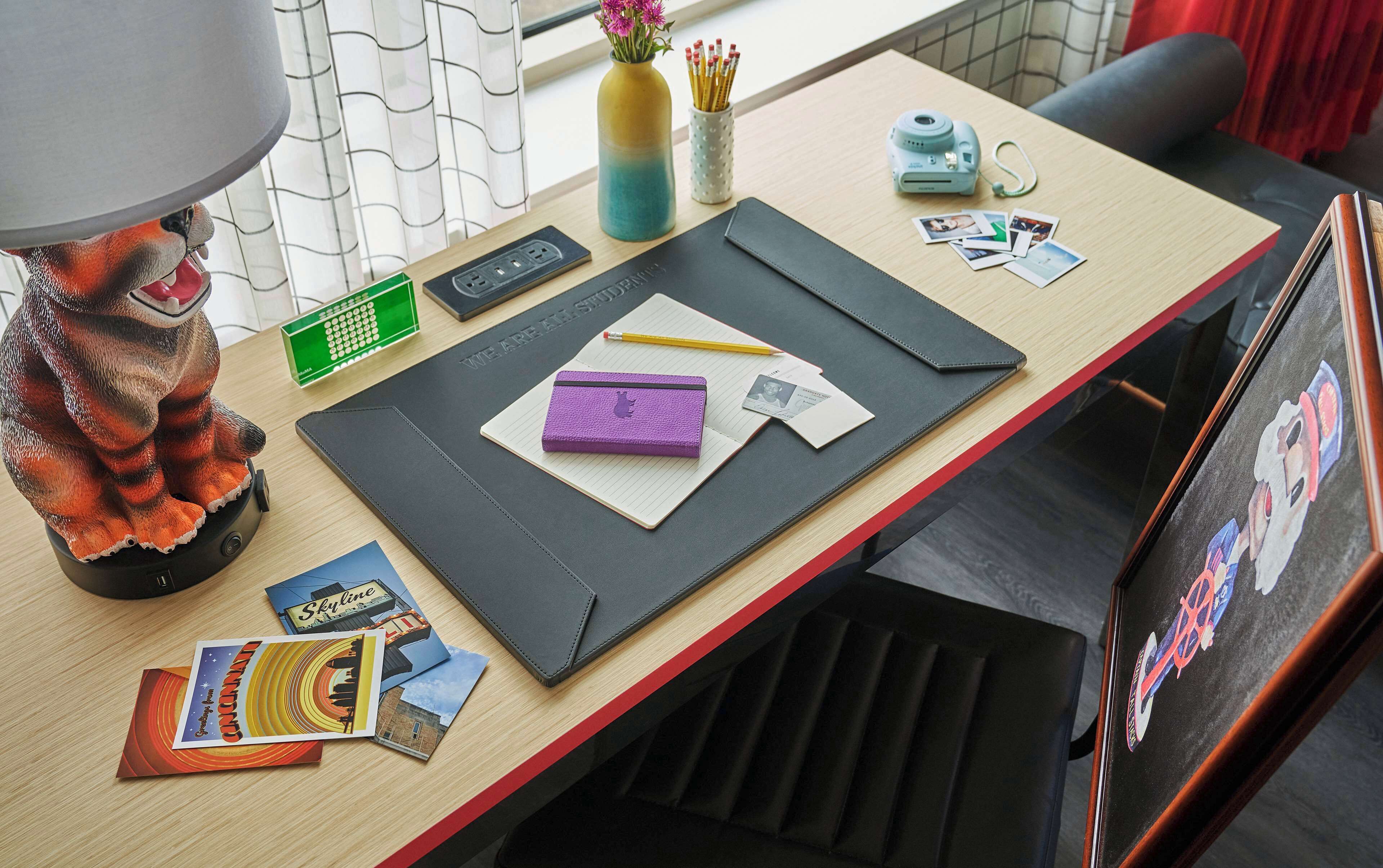 A desk with a lamp, a vase with flowers, a camera, a notebook, a pencil, a purple pencil case, a card, and some postcards.