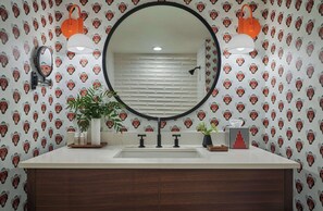 A bathroom with a round mirror, two wall-mounted lights, a sink with a wooden cabinet, and a patterned wallpaper.