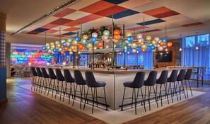 A bar area with a checkered ceiling, colorful hanging lights, and a neon sign in the background.