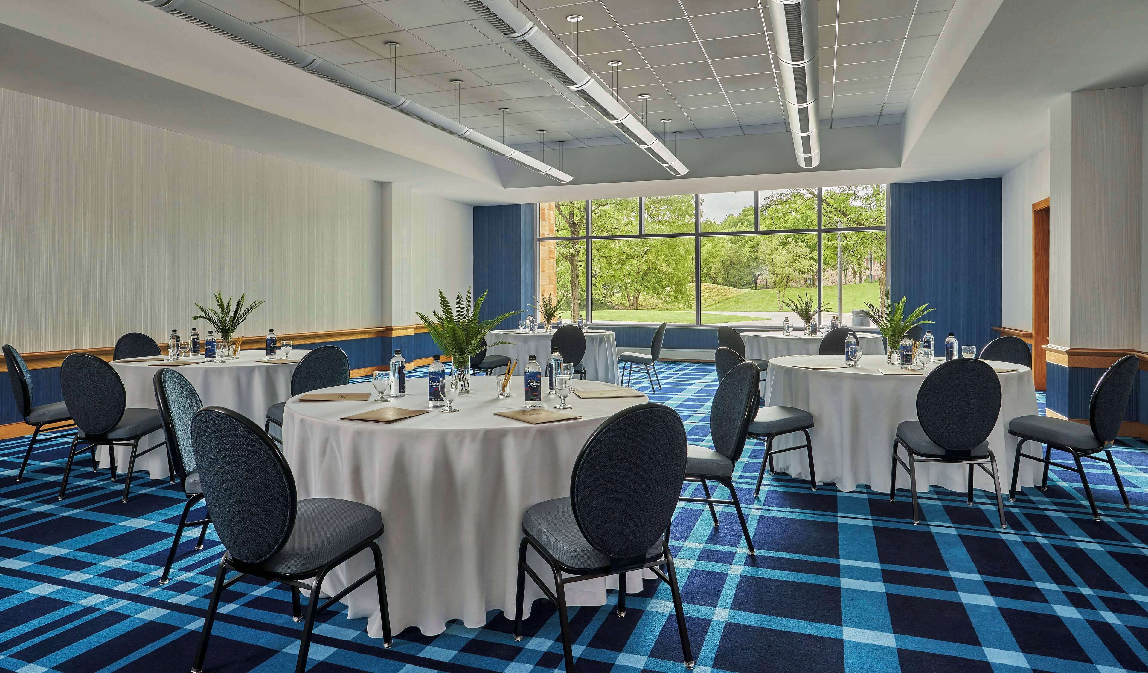 Un salón de conferencias con mesas redondas, sillas y vistas a un paisaje verde a través de amplios ventanales.