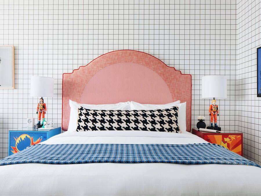 Una cama bien hecha con una colcha a cuadros, un cabecero tapizado en rosa y dos mesitas de noche con lámparas y figuritas de astronautas.