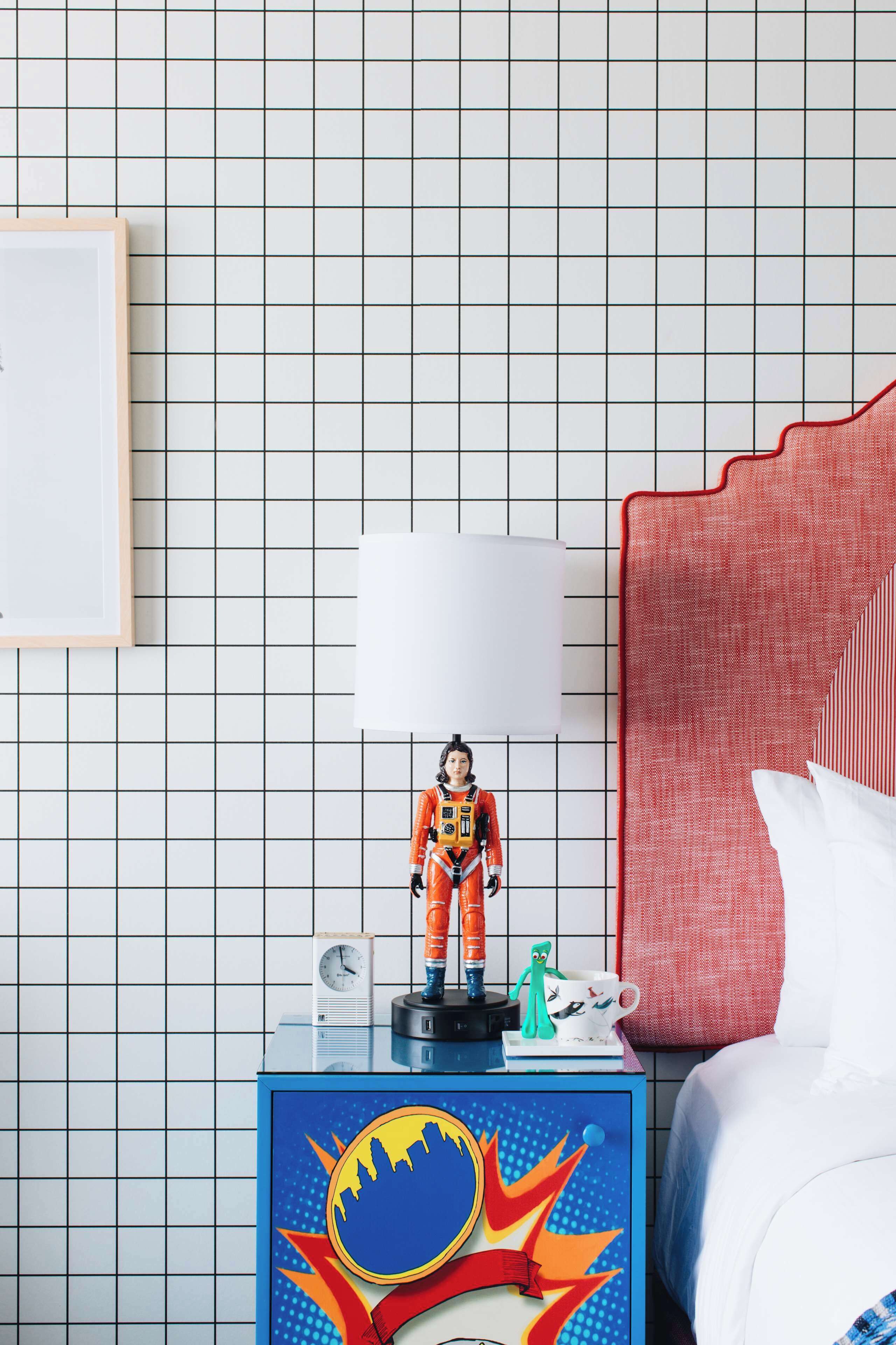 A neatly arranged bedroom with a bed, a nightstand with a clock, a small figurine, and a colorful wall decoration.