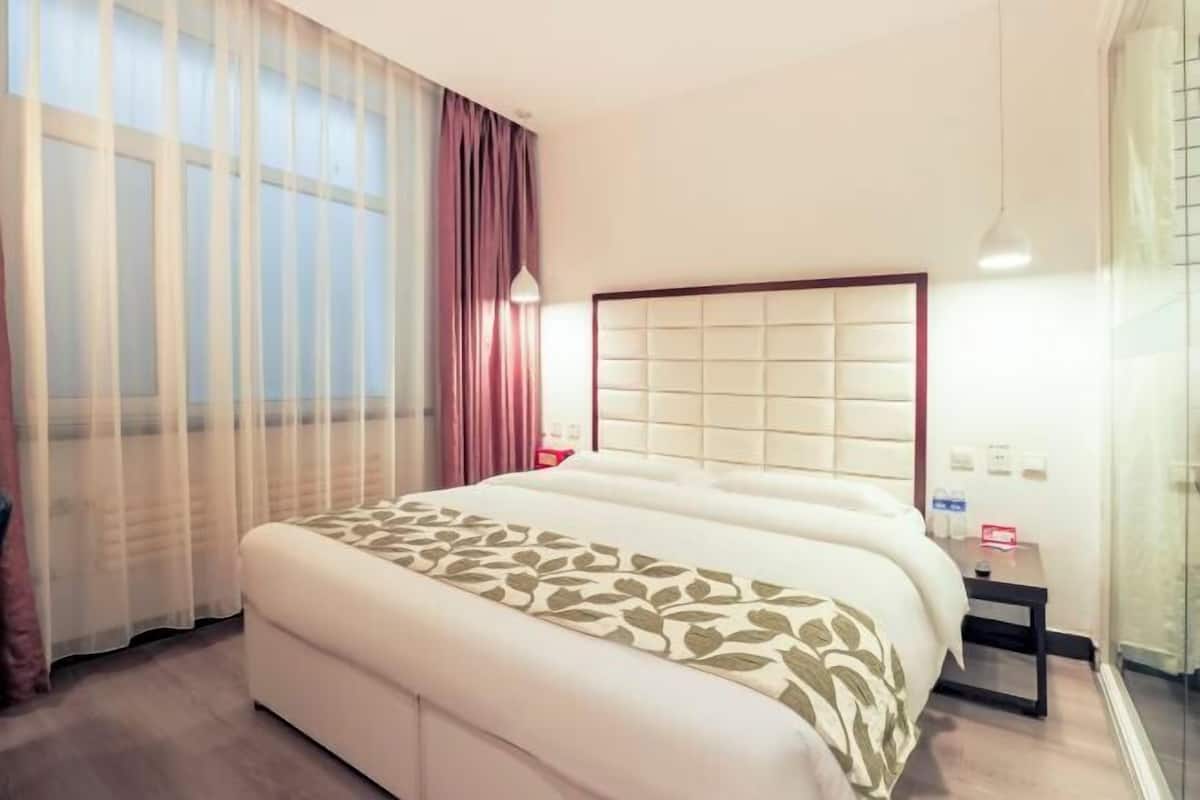 A hotel room with a large bed, a bedside table, a nightstand, a window with sheer curtains, and a wall-mounted light fixture.